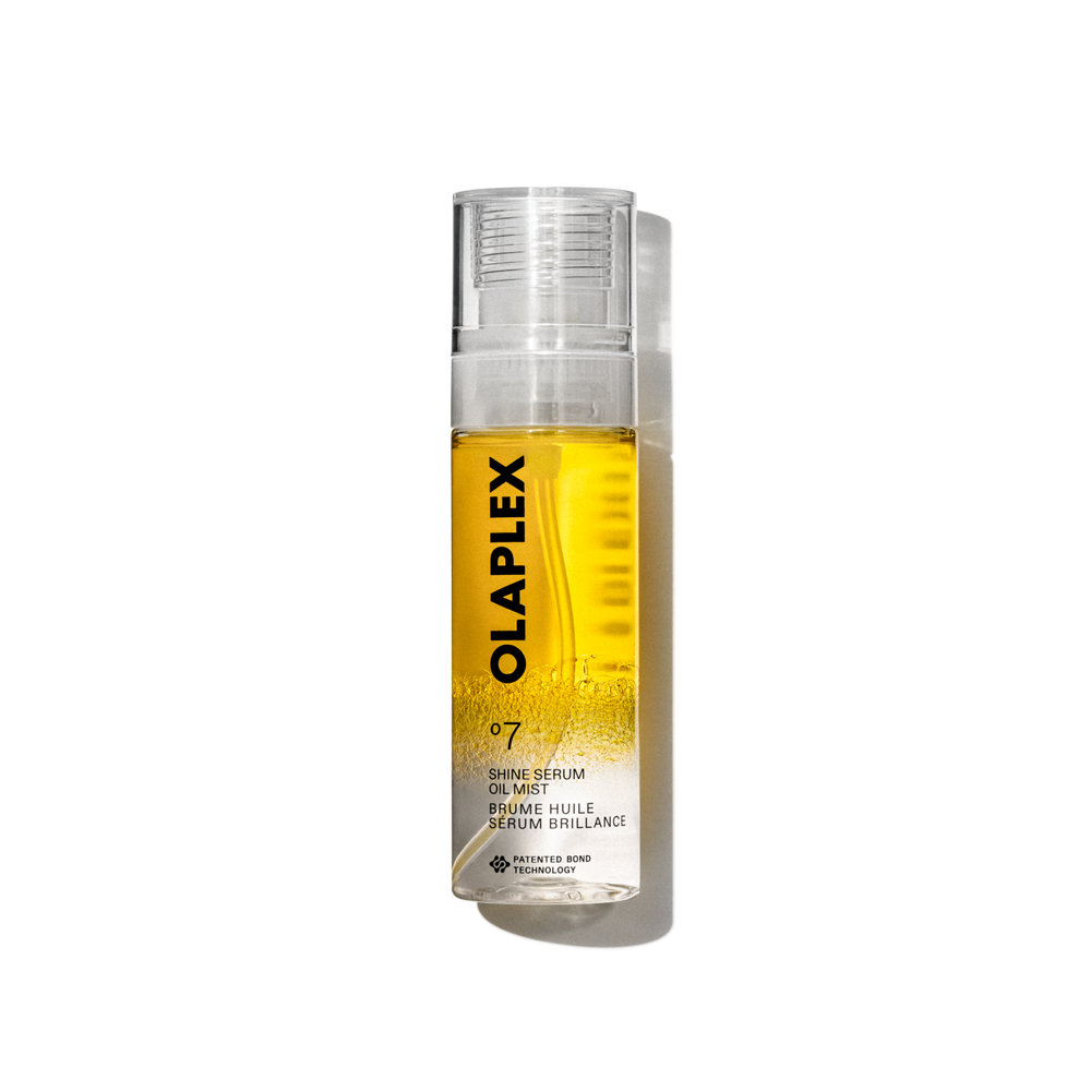 A product packshot of Olaplex No. 7 Shine Serum Oil Mist in an 80ml clear spray bottle. The bottle features a bright yellow liquid visible inside, a transparent cap, and a clean white label at the bottom with black text displaying the product name, details, and the signature "No. 7" logo. The bottle is isolated against a transparent grid background. - main view
